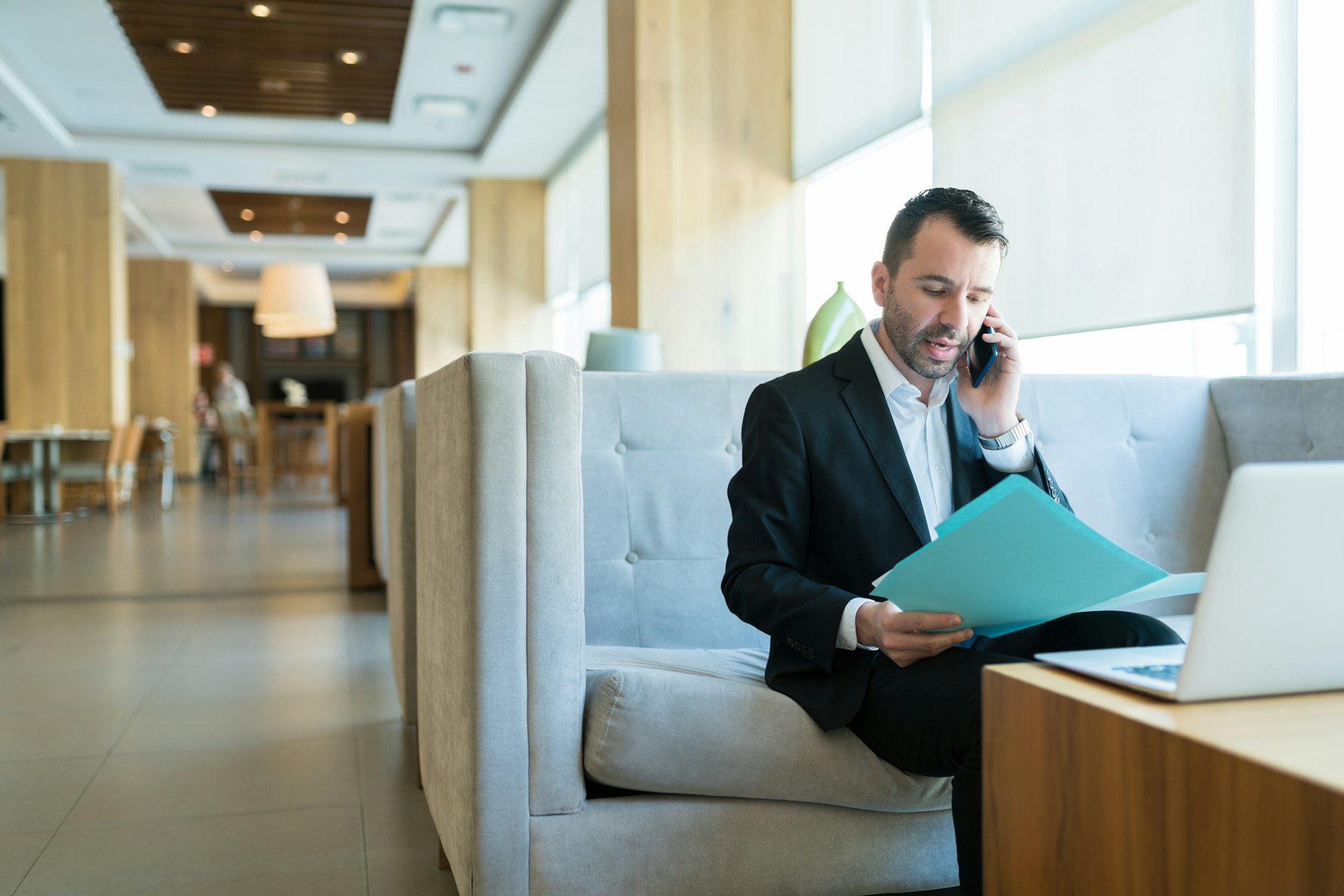 Man in business attire sitting on a lounge in a modern office lobby, speaking on the phone while reviewing documents and using a laptop, representing executive travel and professional planning.