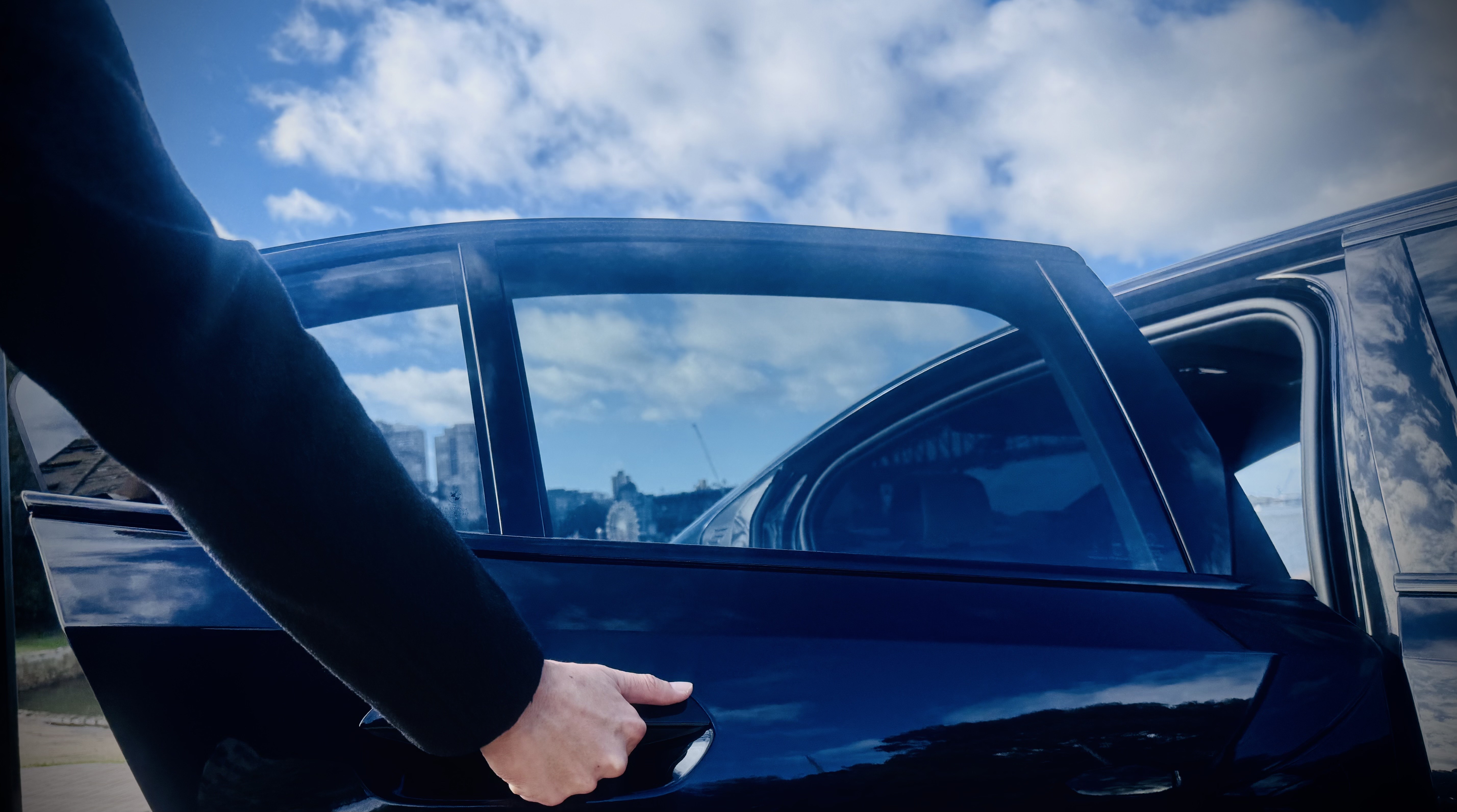 Evoke chauffeur opening the rear passenger door of a BMW i5 executive sedan, ready to welcome a client.