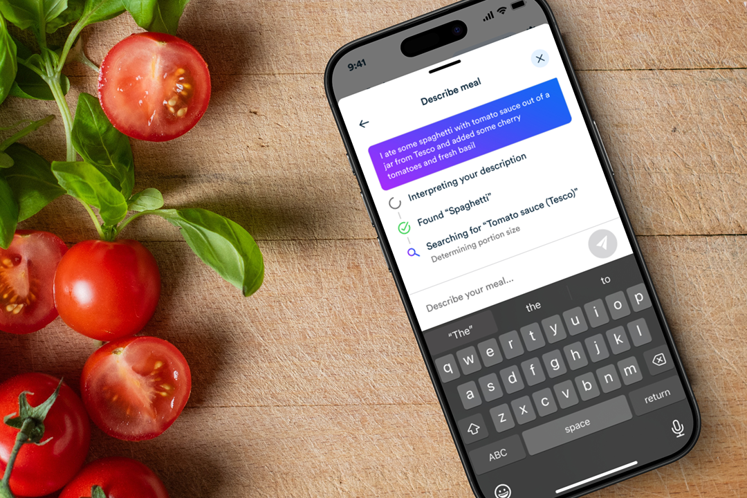 Smartphone on a wooden surface displaying a meal description app with text about spaghetti and tomato sauce, surrounded by fresh tomatoes and basil leaves.