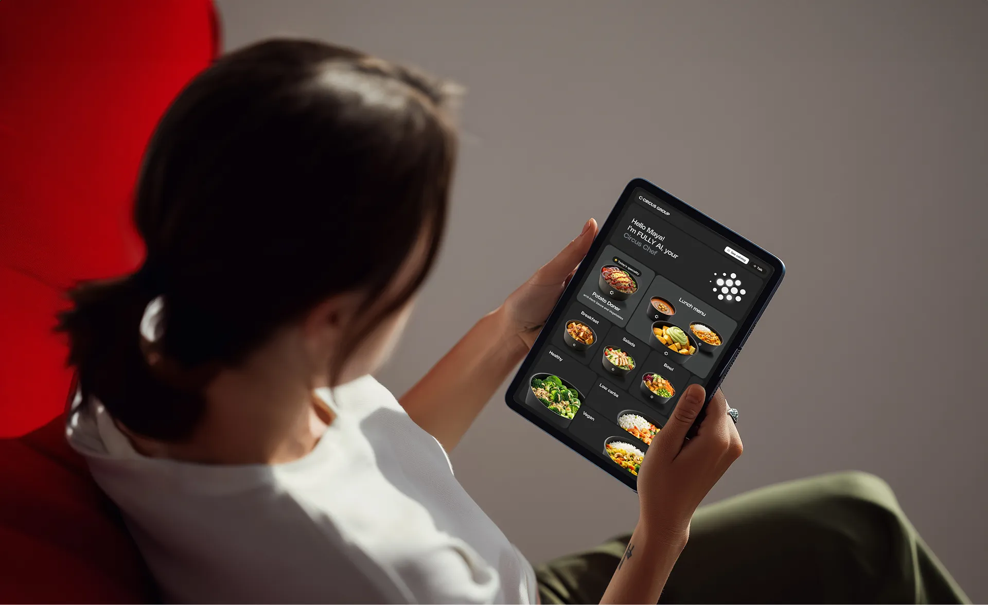 Person sitting on a red chair holding a tablet displaying the Circus Group chef interface with a variety of food menu categories including vegan, low-carb, bowls, pasta, and breakfast.