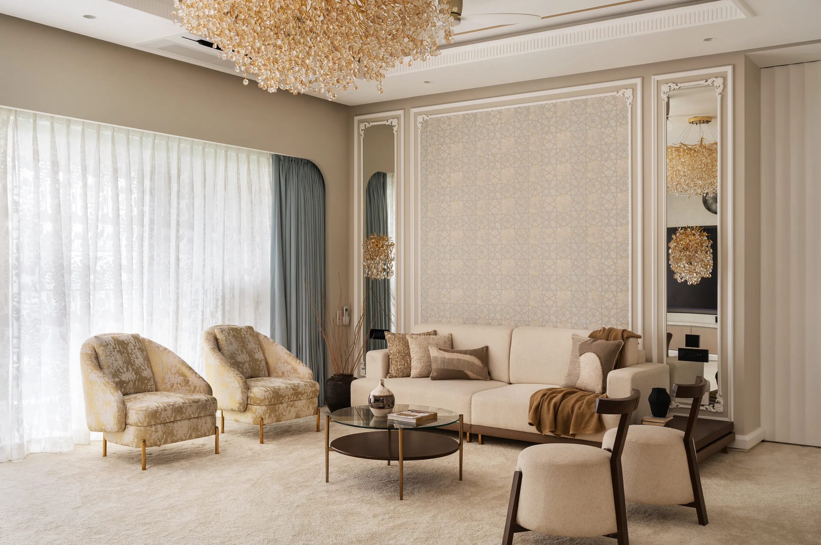 Elegant living room with beige sofa, patterned armchairs, round glass coffee table, decorative pillows, and a large crystal chandelier.