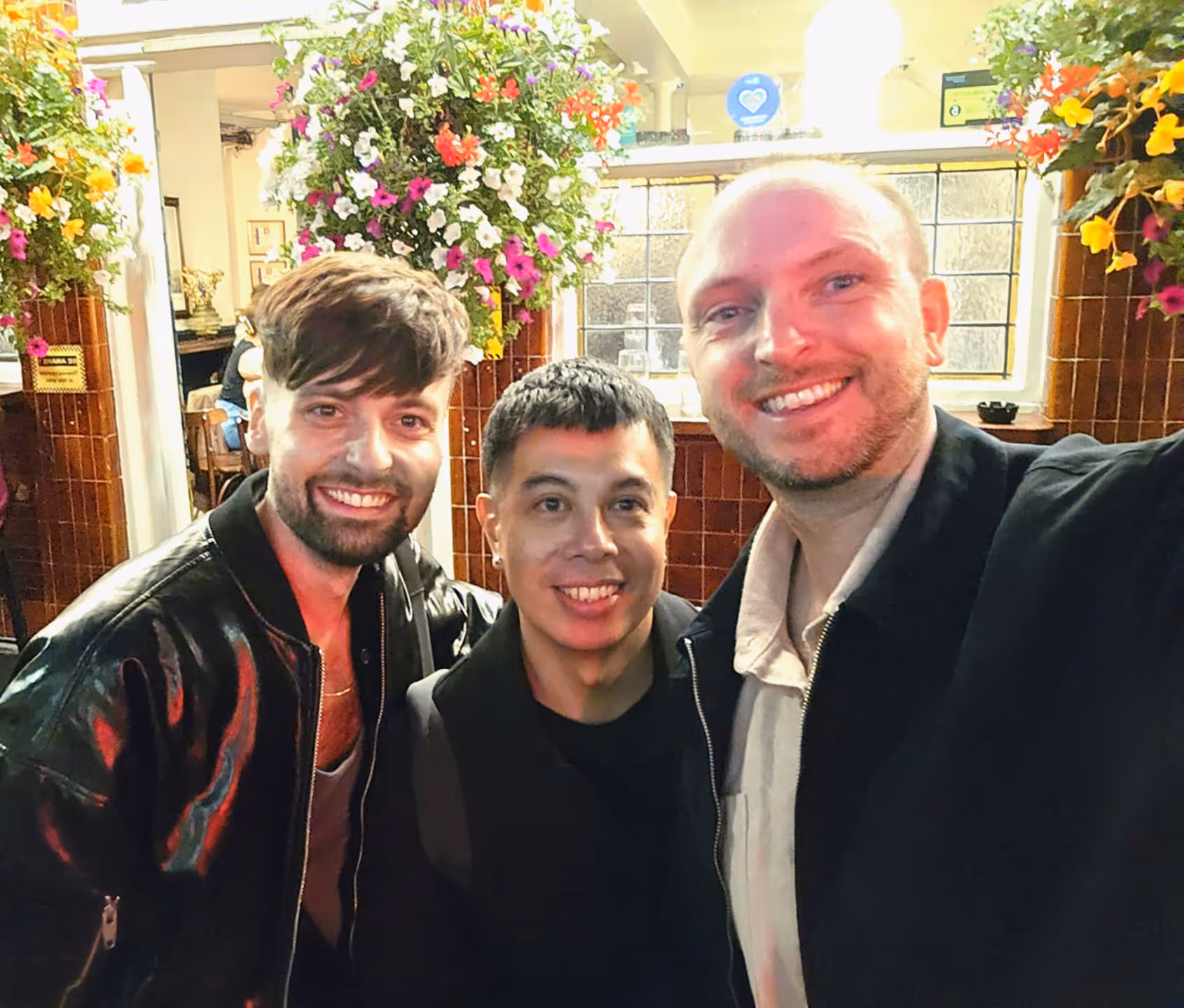 Three smiling men standing close together in a warmly lit indoor space with flowers hanging above.