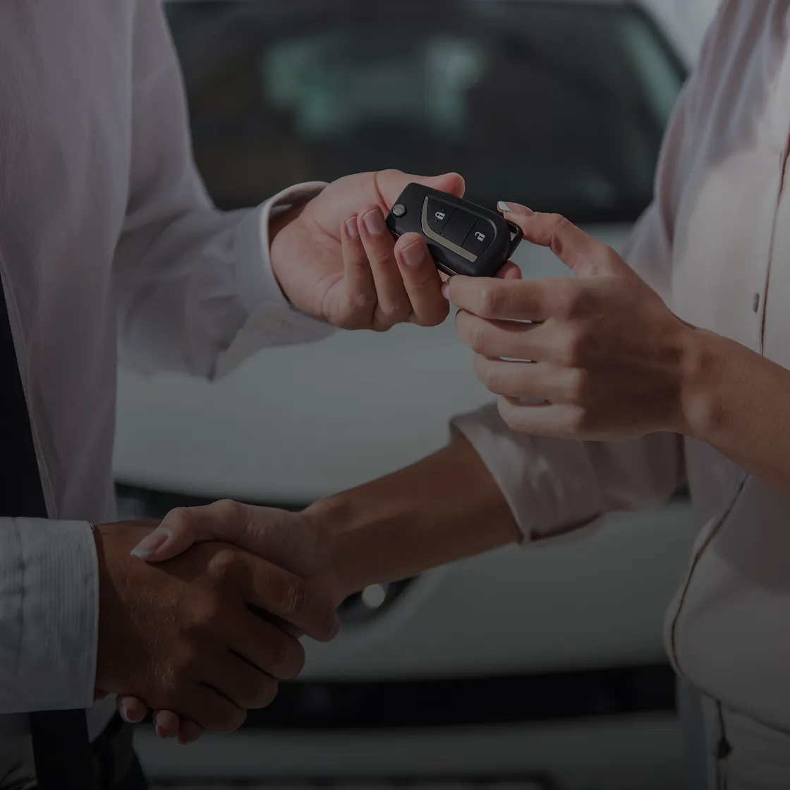 Two people shaking hands while one hands over a car key, with a white car in the background.