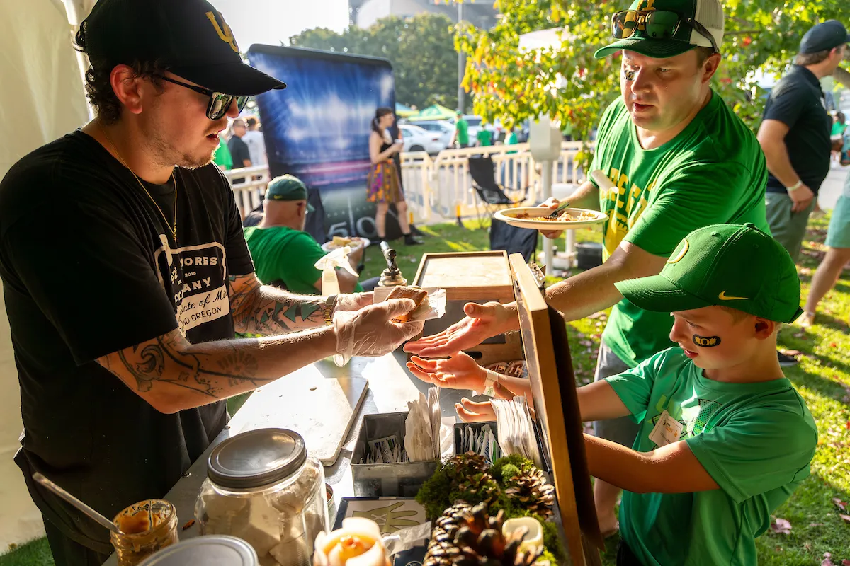 A man and a boy happily receive food from a vibrant food stand in a bustling outdoor setting.