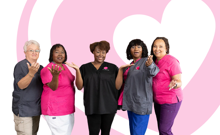 Five diverse healthcare workers in scrubs smiling and gesturing warmly in front of a pink heart background.