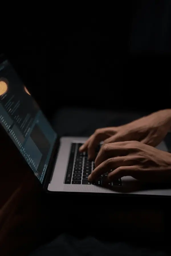 Pair of hands typing on a laptop keyboard with code visible on the screen in a dimly lit environment.