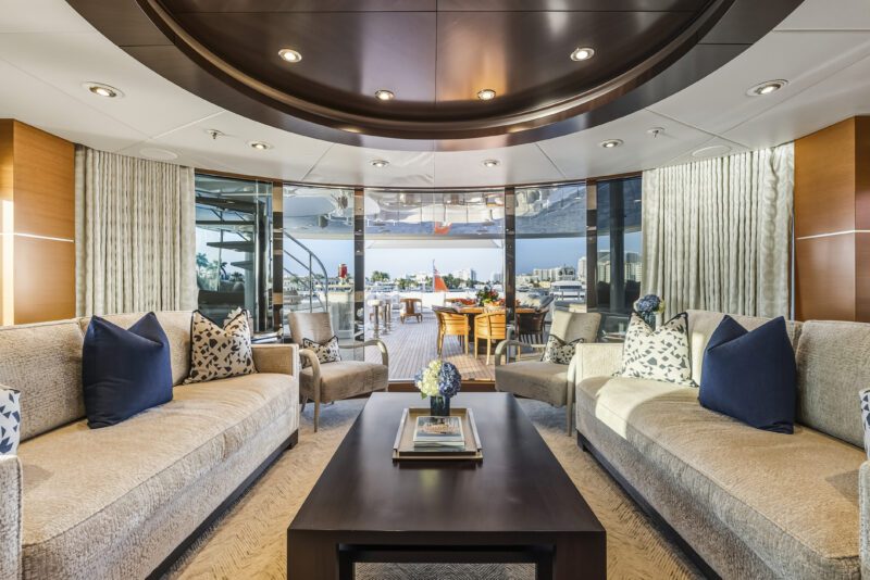 Luxurious Delta Marine Noverra yacht lounge with beige sofas, a central coffee table, and large windows opening to an outdoor dining area with city and water views—ideal for a five-star yacht charter experience.
