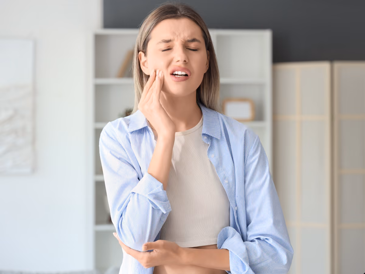 Young woman holding her jaw with a pained expression, illustrating toothache or dental pain while standing indoors in a bright home setting.