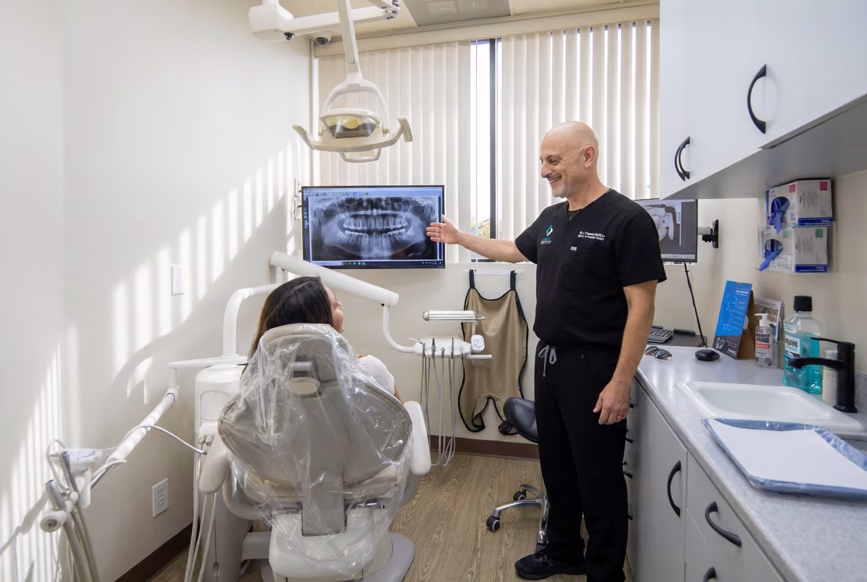 A Clairemont Dentistry dentist smiles while pointing to a panoramic dental X-ray on a monitor for a patient seated in a dental exam chair.
