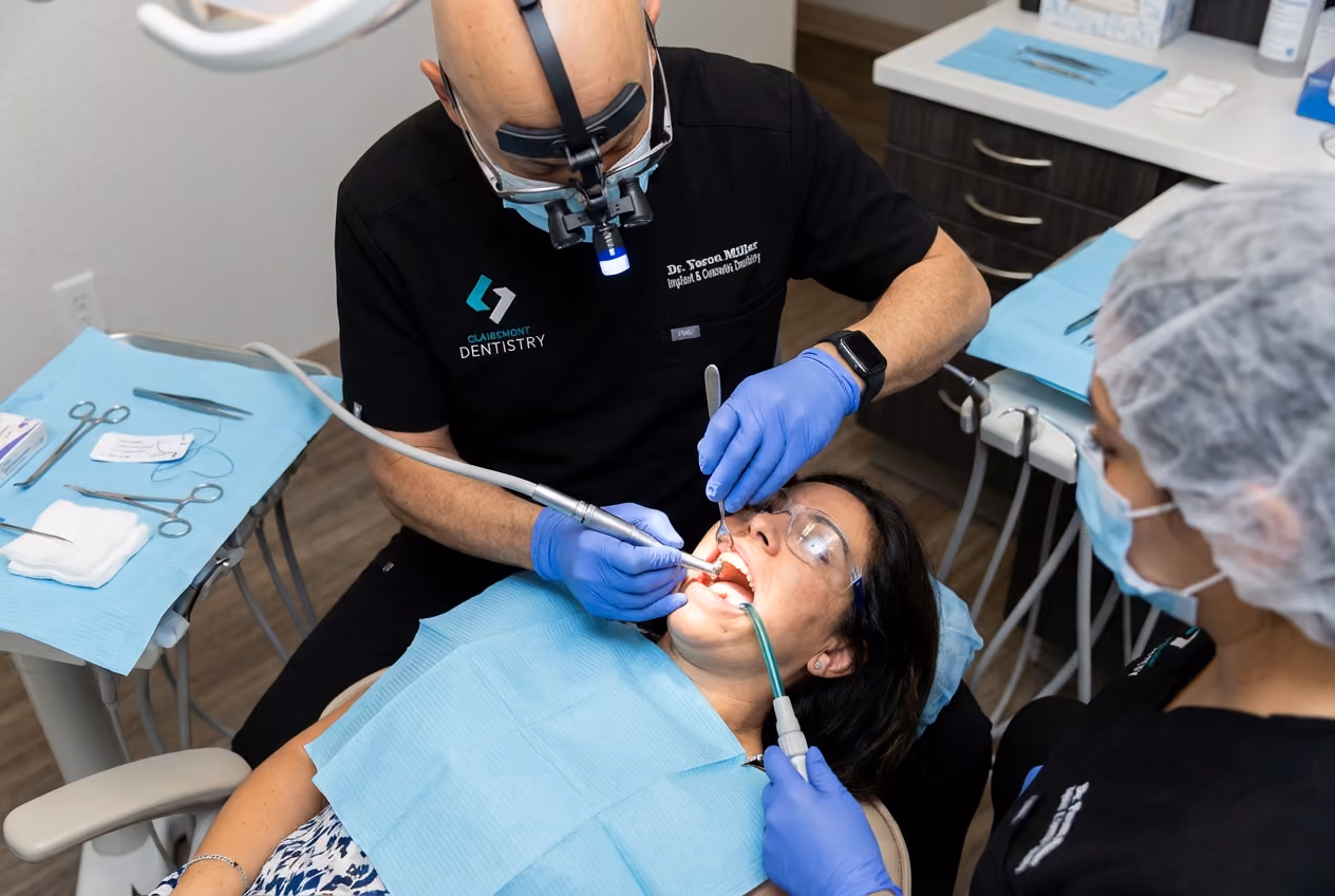 A Clairemont Dentistry dentist wearing magnifying loupes performs a dental procedure on a patient while a dental assistant uses suction in a treatment room with sterile instruments nearby.