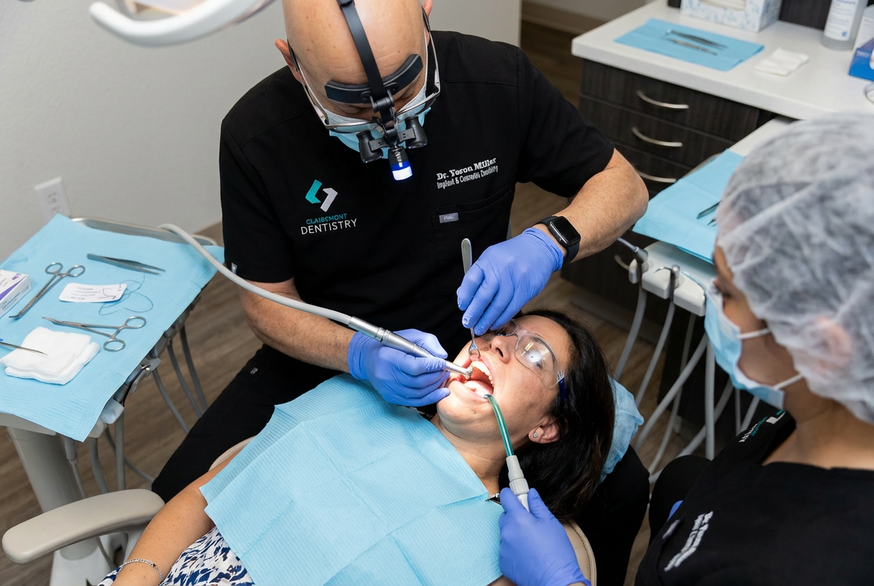 A Clairemont Dentistry dentist wearing magnifying loupes performs a dental procedure on a patient while a dental assistant uses suction in a treatment room with sterile instruments nearby.