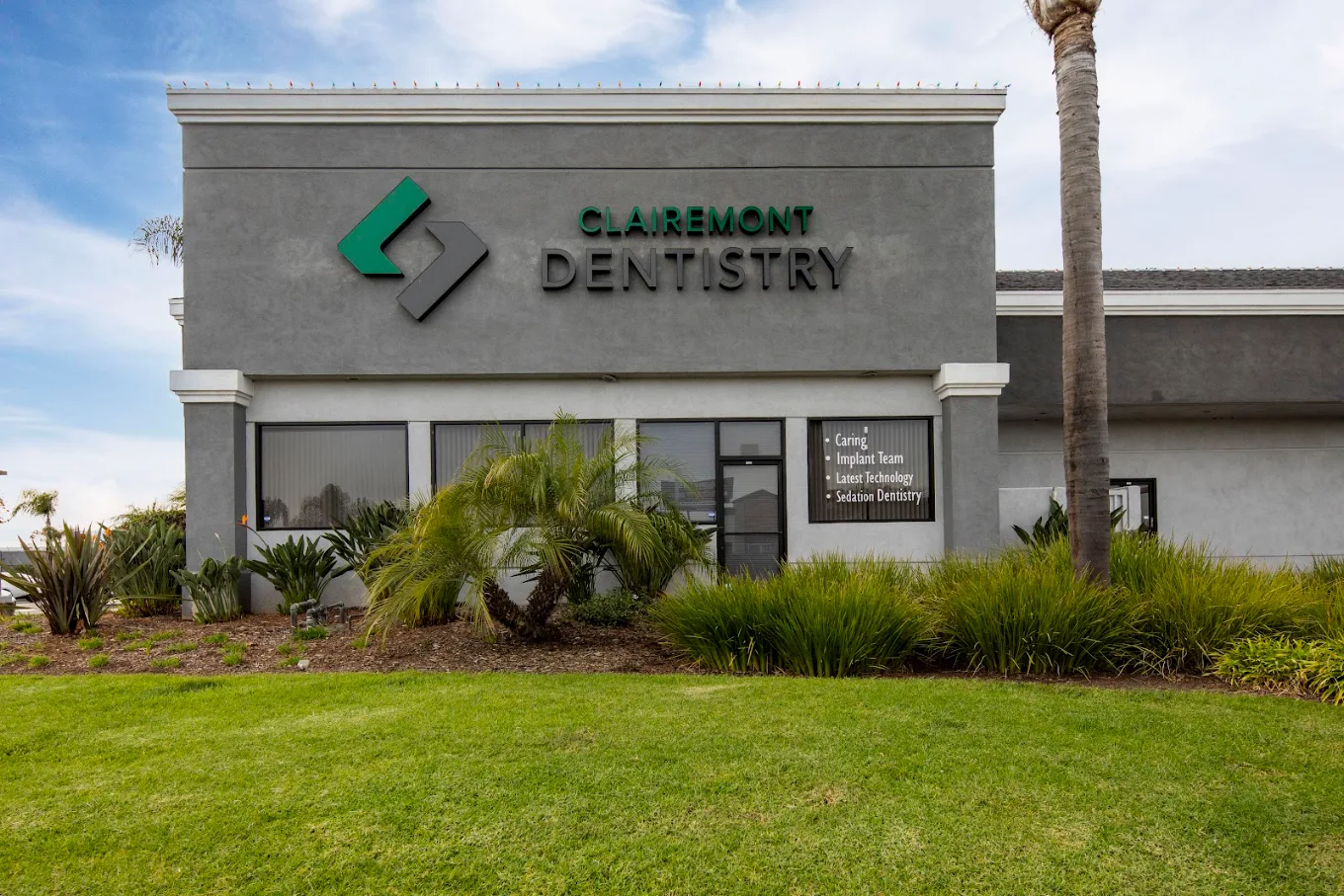 A modern two-story dental office building with large glass windows and "CLAIREMONT DENTISTRY" on the sign, with a man in a white lab coat exiting, and palm trees and blurred cars and people on the street.