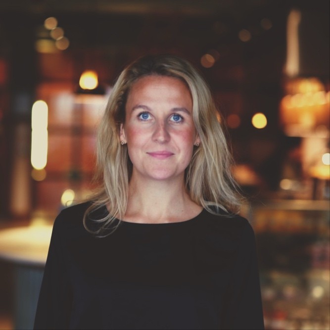 Portrait of a Kimilia wearing a black top, standing indoors with blurred warm lighting in the background.