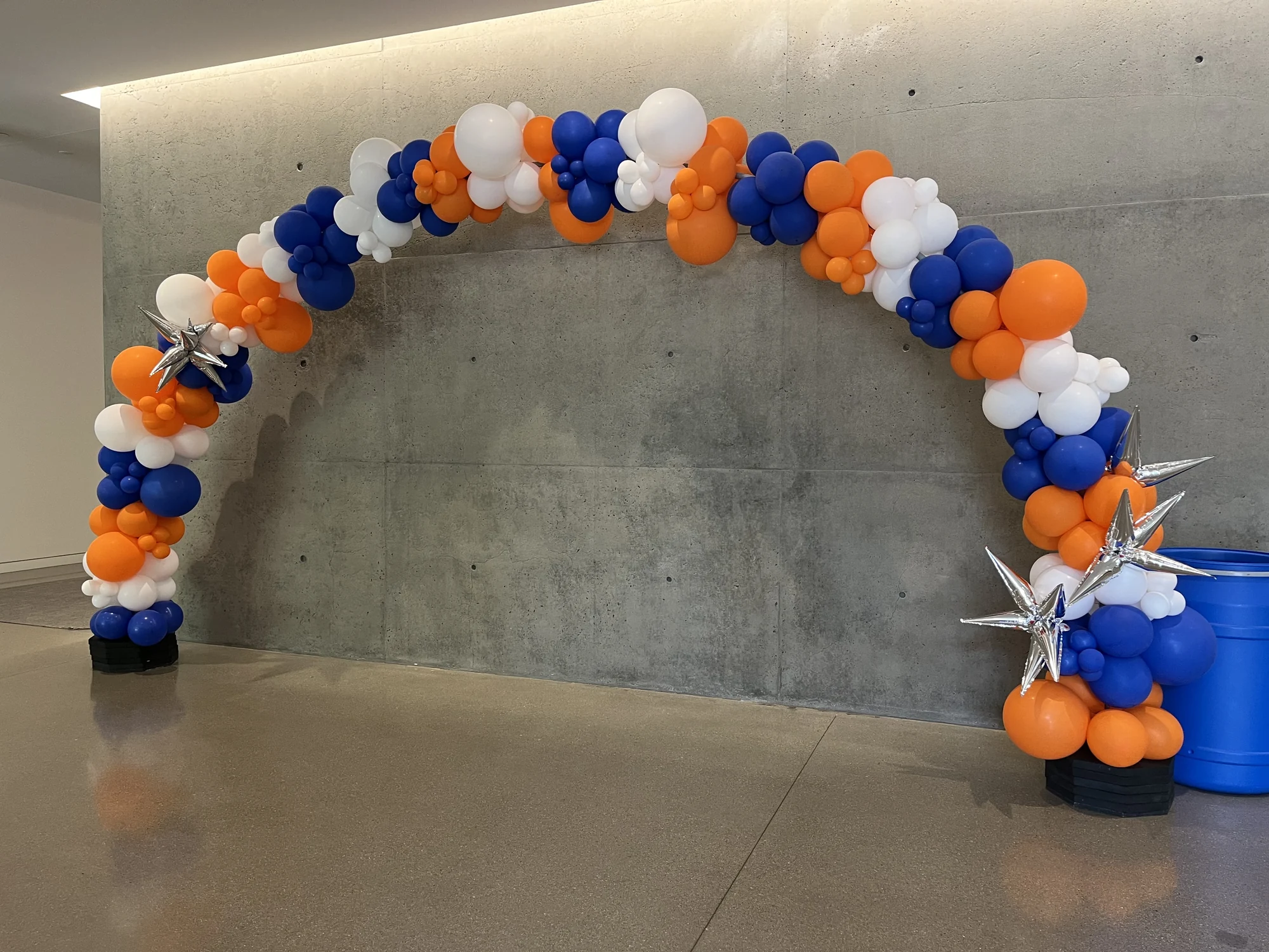 Blue, orange and white balloon arch with silver star accents against a concrete wall