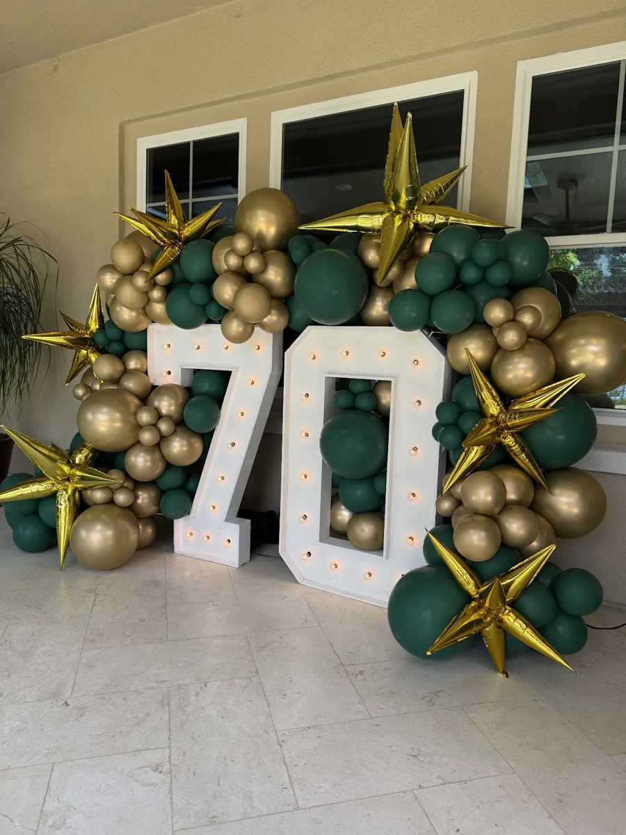 Forest green and gold balloon installation around illuminated 70 marquee numbers.