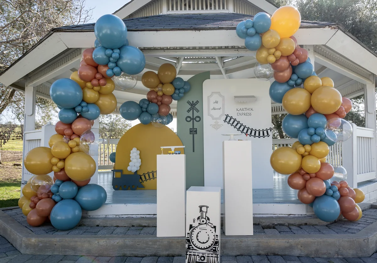Blue, mustard, and rust balloon garlands framing a train-themed birthday backdrop in a gazebo.