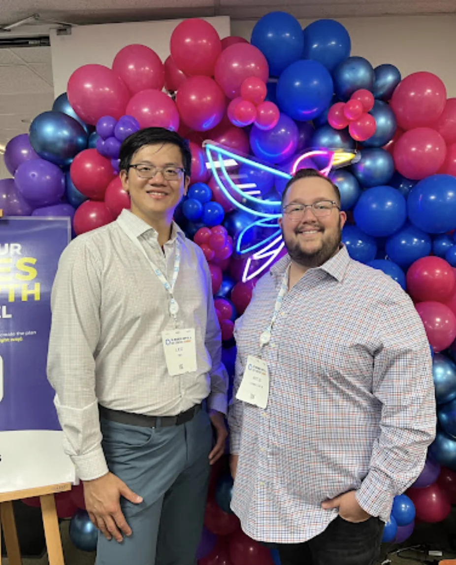 Two guests in front of a pink, blue, and purple balloon wall at a corporate event.