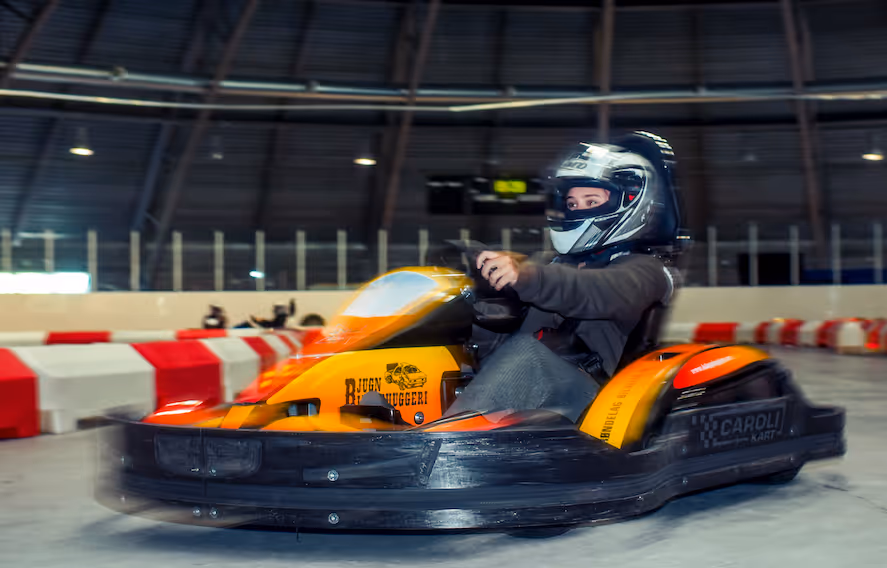 Person med hjelm kjører en oransje go-kart på innendørs bane omgitt av røde og hvite barriereblokker.