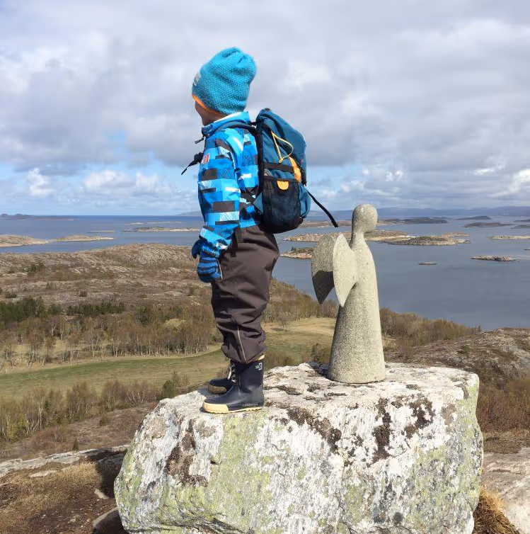 En gutt i blå lue og jakke med ryggsekk står på en stein ved siden av en engelstatue, med utsikt over kystlandskap og vann.