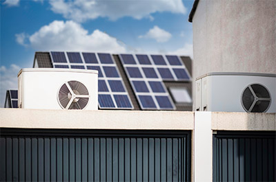 Zwei Wärmepumpen stehen auf einem Garagendach und im Hintergrund ist ein Hausdach, auf dem mehrere Solarpanels angebracht sind.