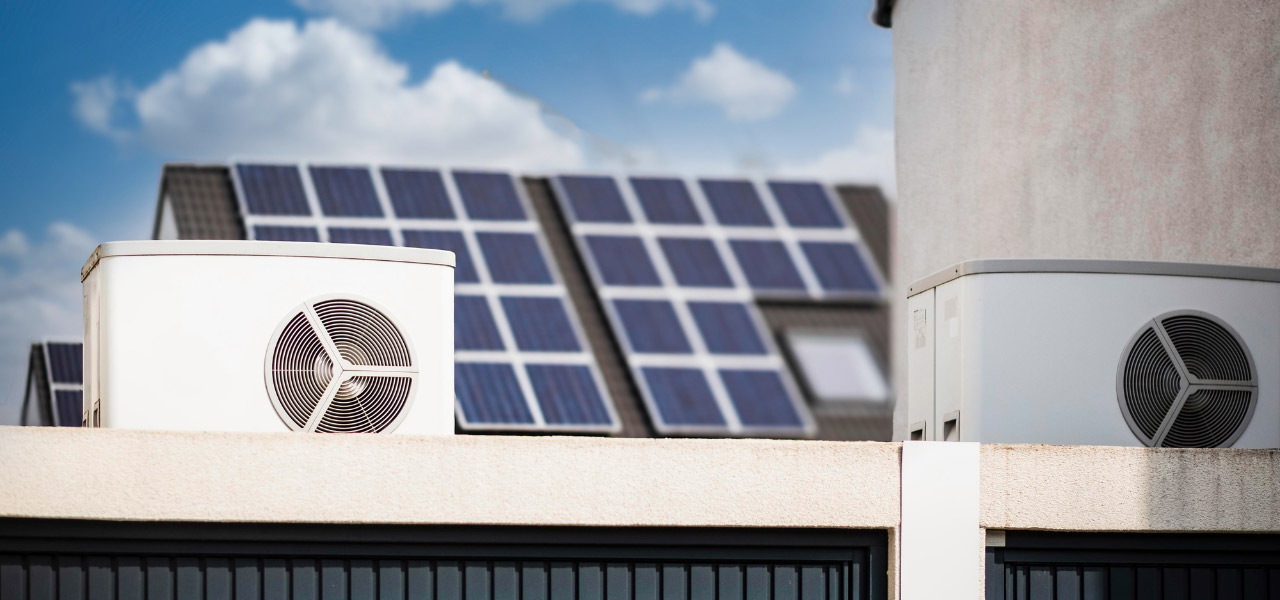 Zwei Wärmepumpen stehen auf einem Garagendach und im Hintergrund ist ein Hausdach, auf dem mehrere Solarpanels angebracht sind.