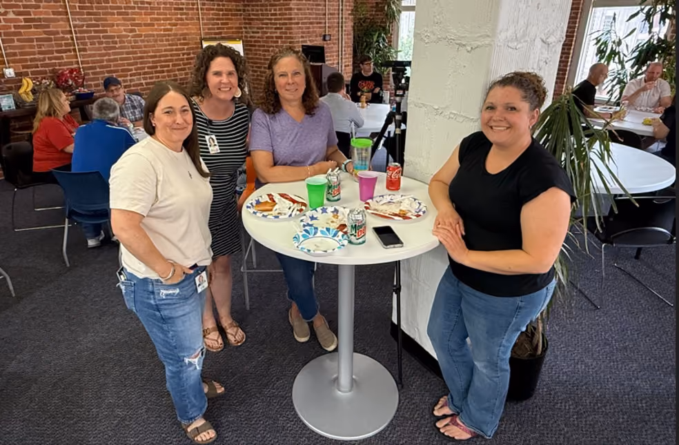 Four women standing around a small round table with drinks and plates in an office break room.
