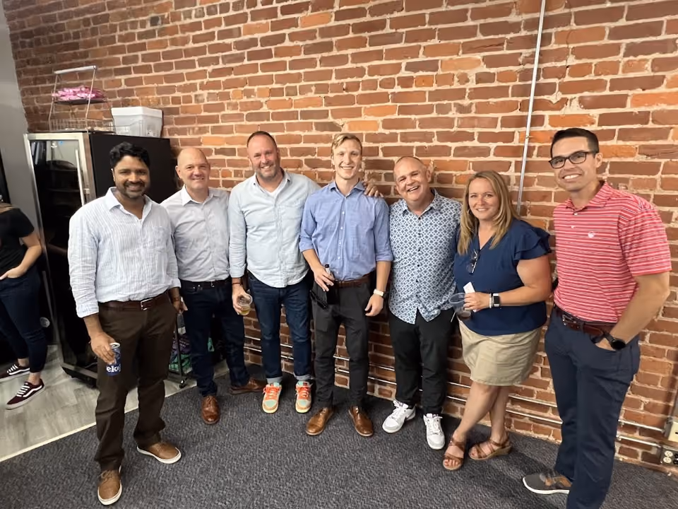 Seven adults smiling and standing side by side against a brick wall in a casual office setting.
