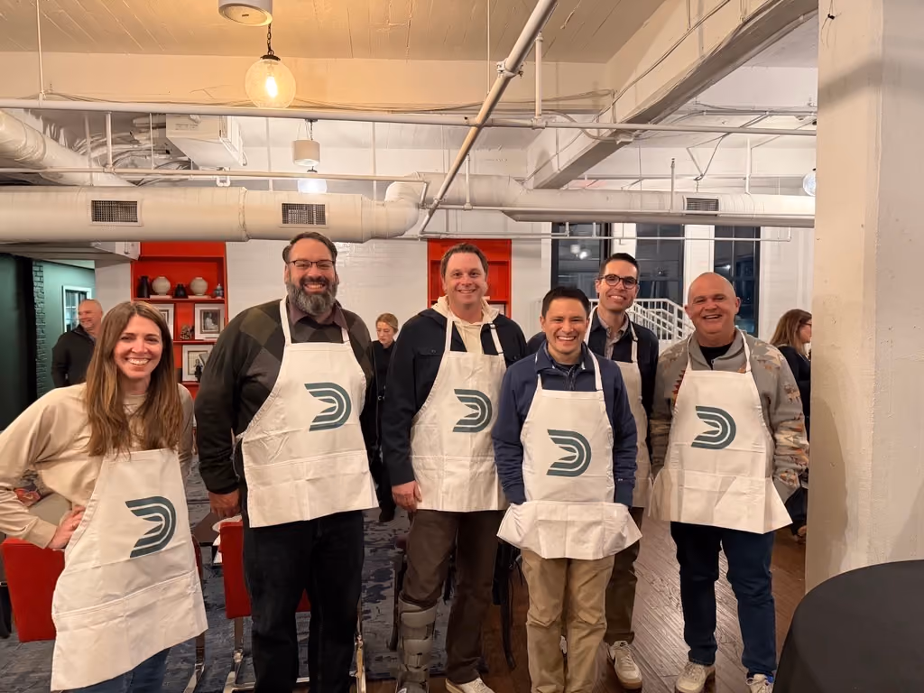Six people standing indoors wearing aprons with a logo, smiling at the camera.