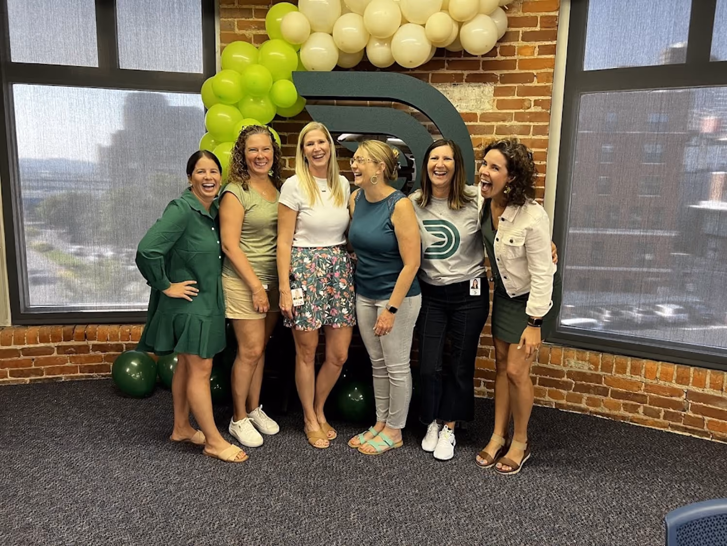 Six women standing indoors in front of green and white balloon arch and company logo, smiling and laughing.