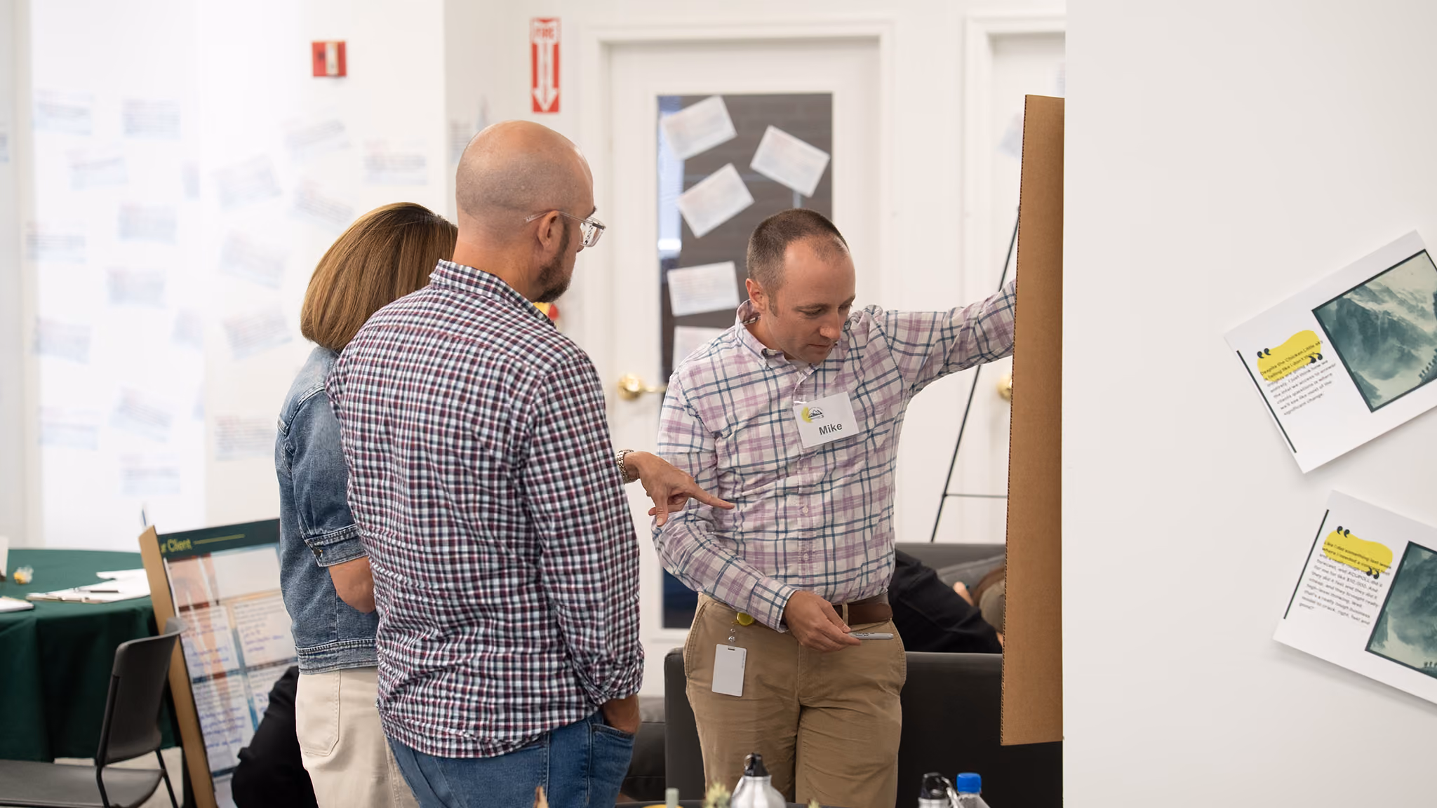 Three people in an office reviewing a board presentation; man on right wearing a name tag reading Mike.