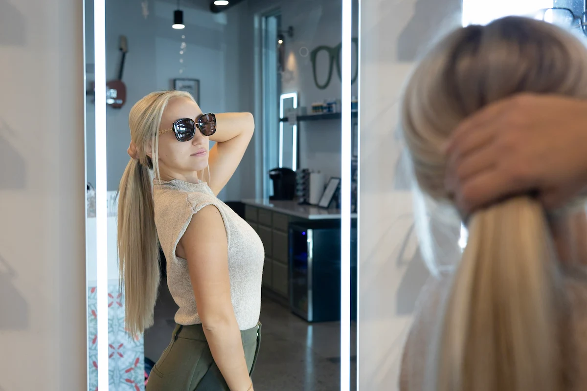 Woman with long blonde hair wearing dark sunglasses looks at herself in a lighted mirror while holding her ponytail.