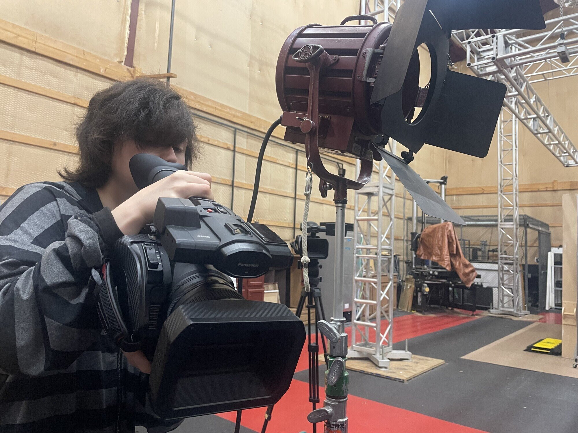 A photo of a young man with dark hair looking into the eyepiece of a video camera with lighting and rigging equipment visible in the background.