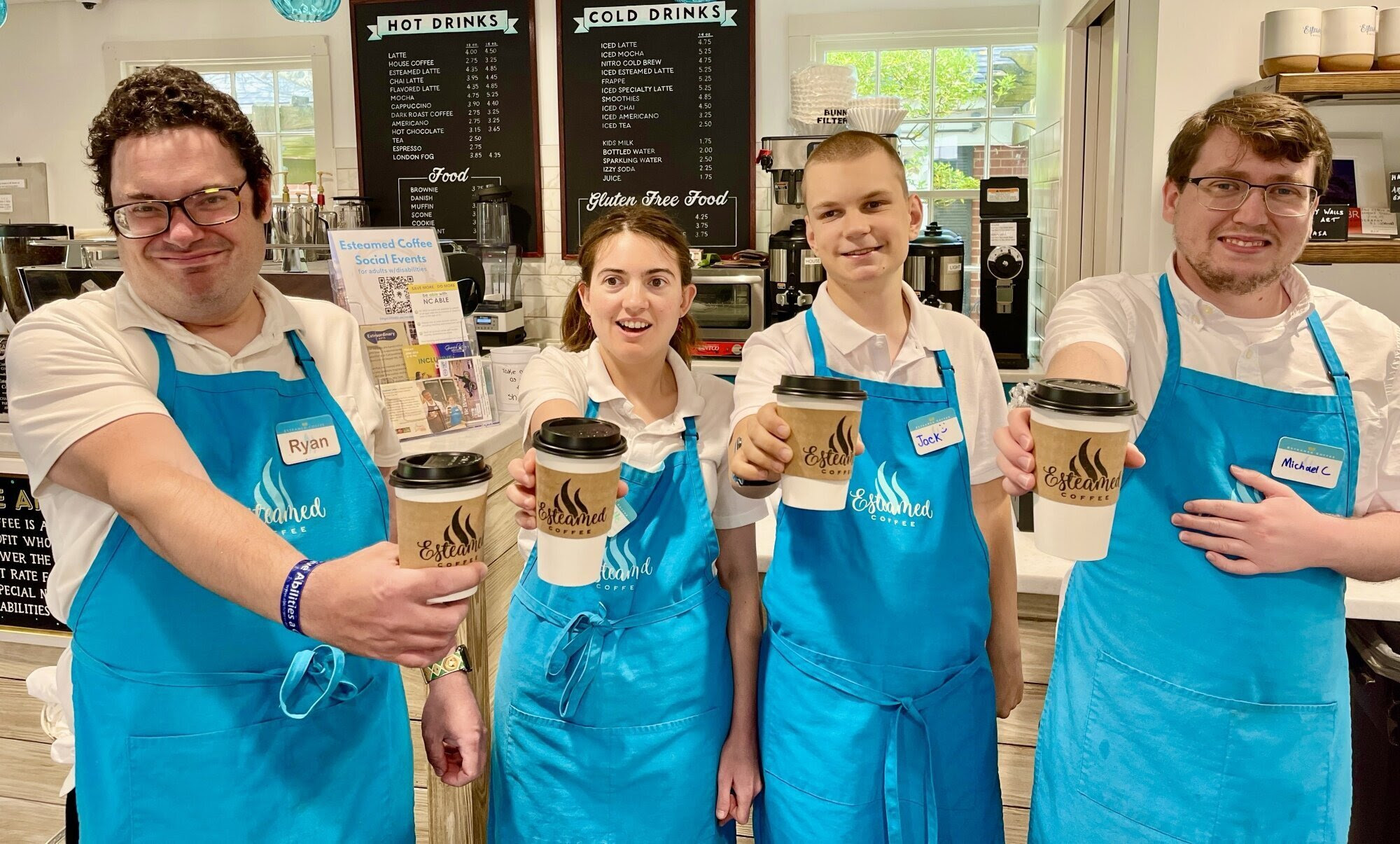 Four coffee shop workers in blue aprons smiling and holding to-go cups out toward the camera.