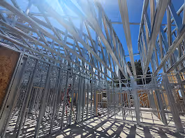 Steel frame structure under construction on a concrete foundation with a person working on a ladder against a partly cloudy blue sky.