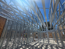 Steel frame structure under construction on a concrete foundation with a person working on a ladder against a partly cloudy blue sky.