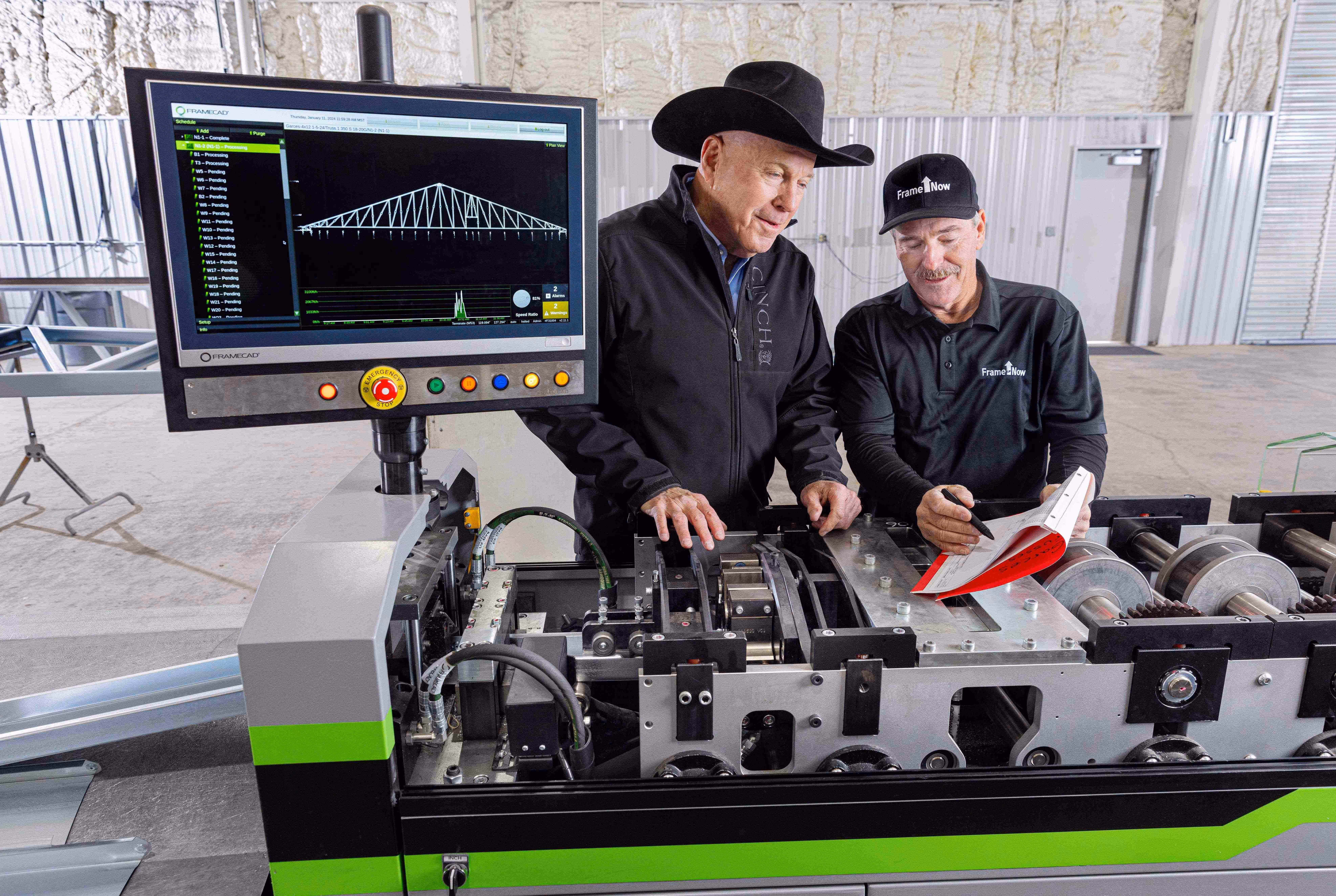 Two men in black clothing and hats examining a high-tech machine with a computer screen showing a bridge design.