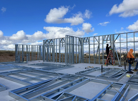Title: Cold-formed steel wall frames rising on a concrete slab at a Red Hawk at J-6 Ranch homesite, with framers, a ladder, and mountains beyond - Description: Cold-formed steel wall frames rising on a concrete slab at a Red Hawk at J-6 Ranch homesite, with framers, a ladder, and mountains beyond