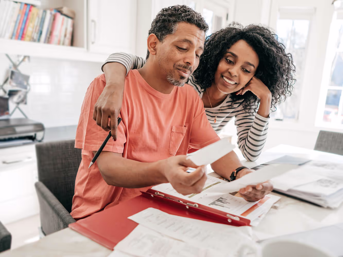 A smiling woman has her arm around a man in a peach-colored T-shirt as they review financial documents while seated at a table