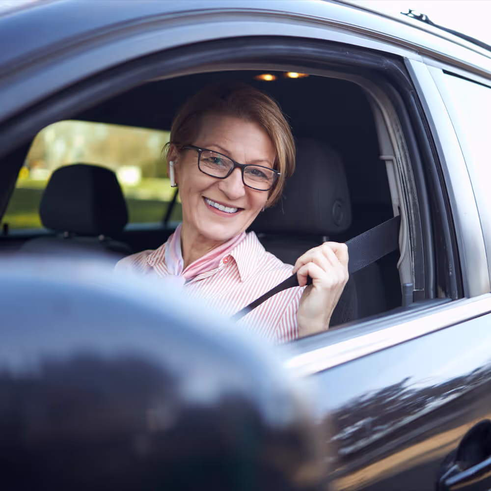 A smiling middle-aged woman in a car putting on her seat belt