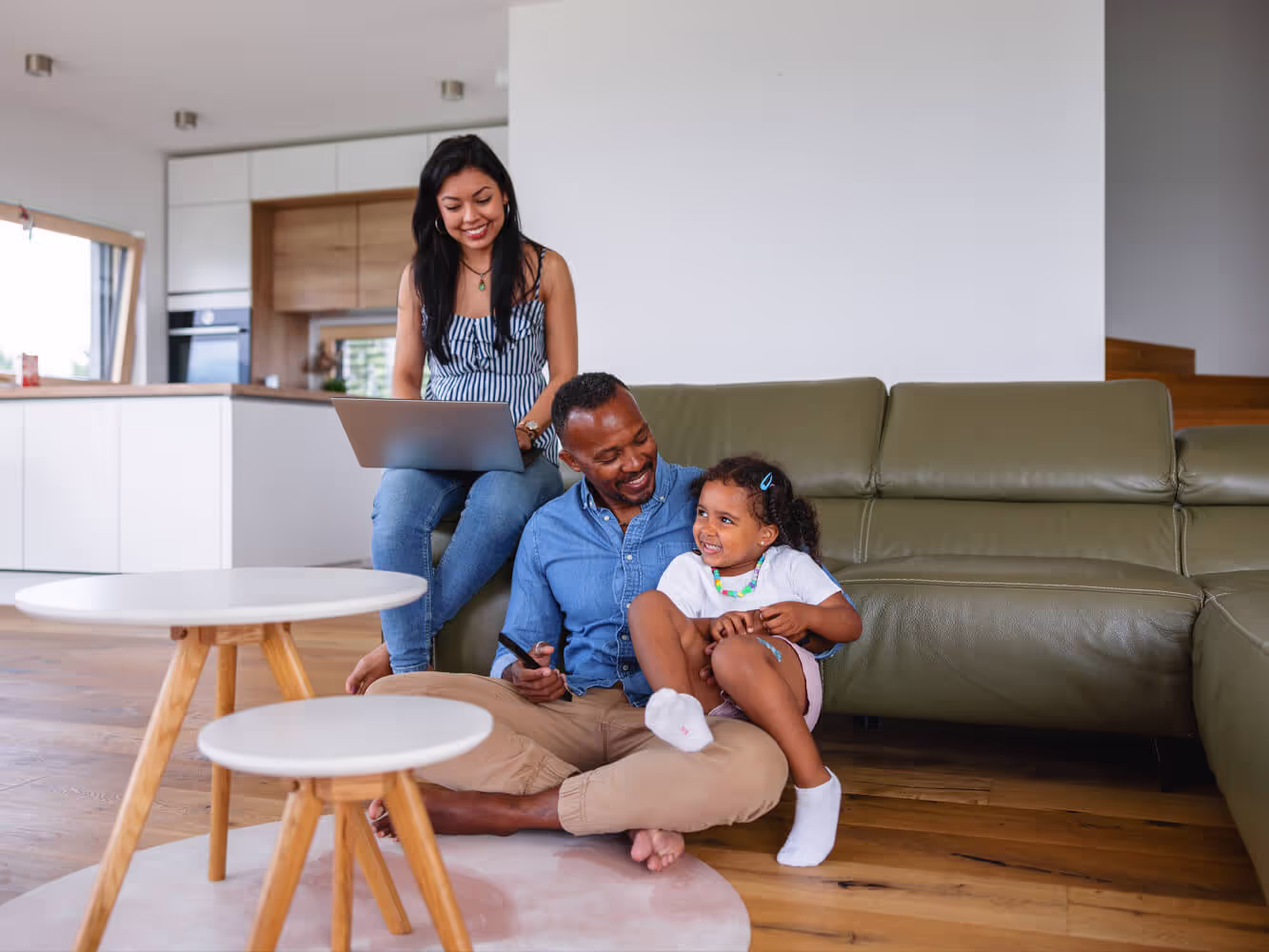 Father and young daughter sitting on the living room floor next to a sofa, while a woman sits on sofa arm holding a laptop