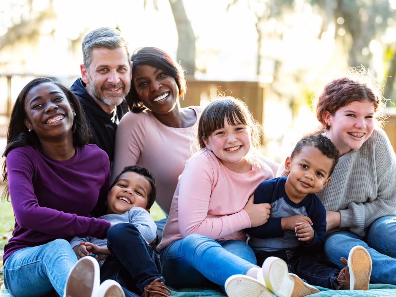 A blended family of man, woman and 5 kids sitting on a blanket on the ground outside