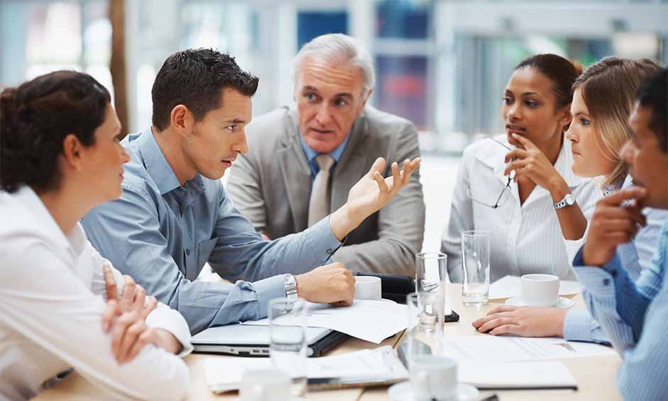 A diverse group of six business professionals engaged in a discussion around a table with documents and glasses of water.