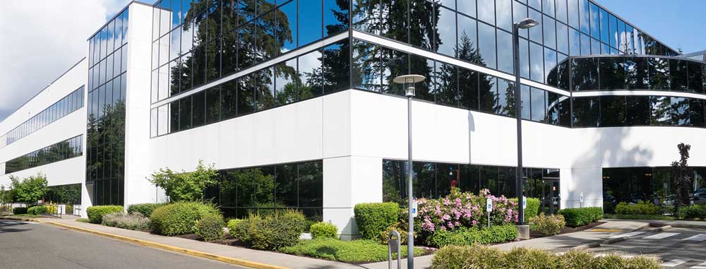 Modern office building with reflective glass windows, surrounded by landscaped bushes and trees under a partly cloudy sky.