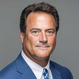 Middle-aged man with dark hair wearing a blue suit and striped tie, looking at the camera against a gray background.