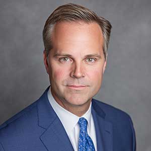 Portrait of a man in a blue suit, white shirt, and blue patterned tie against a gray background.