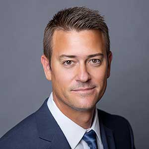 Professional headshot of a man with short brown hair wearing a navy suit jacket, white shirt, and blue tie against a gray background.