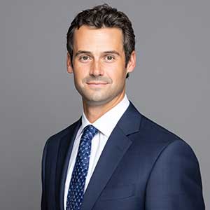 Portrait of a man in a navy suit and patterned blue tie against a gray background.