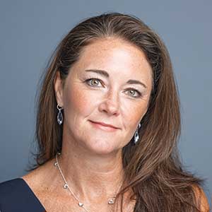 Woman with long brown hair wearing silver earrings and a necklace, against a gray background.