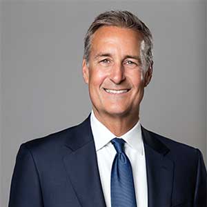 Smiling middle-aged man wearing a blue blazer and checkered shirt against a gray background.
