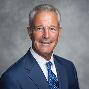 Portrait of a smiling older man with gray hair wearing a navy suit, white shirt, and blue patterned tie against a gray background.